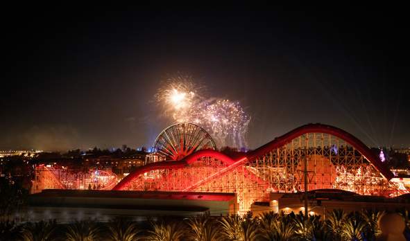Disney fireworks lighting up the night sky over a brightly illuminated roller coaster and Ferris wheel at a theme park in Anaheim, California.