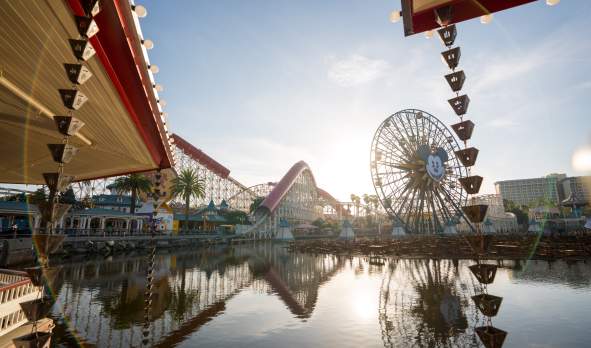 Sunrise at Disney California Adventure Park with a large Ferris wheel and roller coaster, reflected in a serene lake surrounded by palm trees.