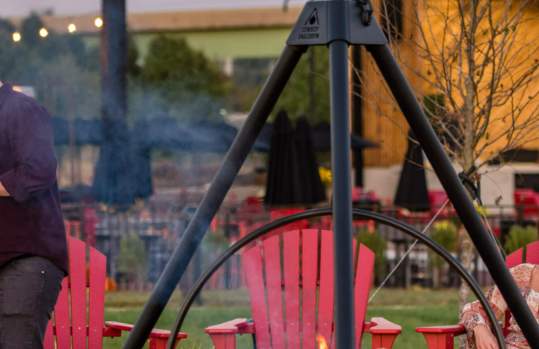 People sitting around a fire in red Adirondack chairs, in the back there is a building and an outdoor stage