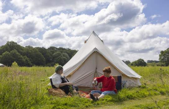 A white canvas tent with two ladies sat in front of it in the grass having a picnic