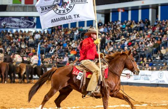 A cowboy wearing a red shirt, tan chaps, and a cowboy hat rides a galloping chestnut horse inside an indoor rodeo arena. He holds a large white flag that reads "West Texas Ranch Rodeo, Amarillo, TX," with a logo in the center. The background shows a packed audience in the stands and several other riders on horseback in the arena. The dirt-covered floor and western attire emphasize the rodeo setting.