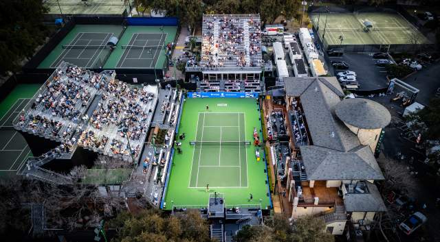 Aerial image of the ATX Open tennis court at Westwood Country Club with a bright green, illuminated court in the middle surrounded by spectator stands with people sitting on bleachers.
