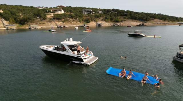 A boat floating on Lake Travis with people hanging out on the boat and on a large floaty in the water.