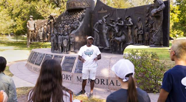 Javiar Wallace, founder of Black Austin Tours, standing in front of a memorial statue, talking to a group of people giving a tour.