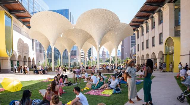 Groups of people standing around and sitting on picnic blankets across the Blanton Museum's lawn, in between museum buildings.