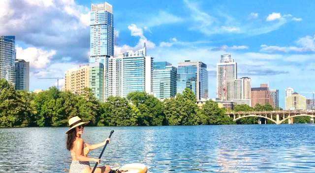 Girl in swimsuit and hat, sitting on paddle board on Lady Bird Lake looking towards the downtown skyline.