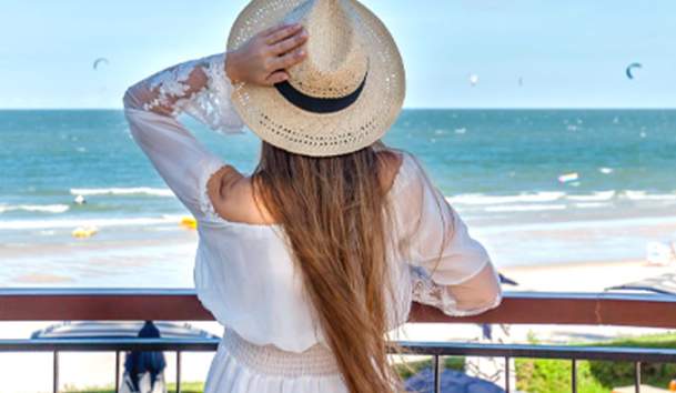 Woman in sunhat, standing on hotel balcony, overlooking the beach and ocean.