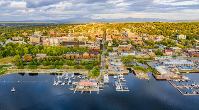 Aerial view of Burlington by a lakeshore with marinas, docks, and numerous buildings. Greenery surrounds the area, and mountains are visible in the background.