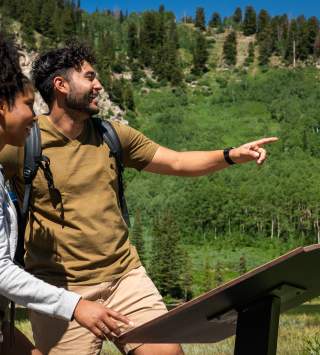 Man and Women with day hike backpacks looking off screen and pointing with a scenic background and information sign in front of them