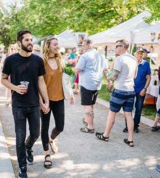 Couple strolling down the sidewalk in a crowd in front of several farmer's market booths