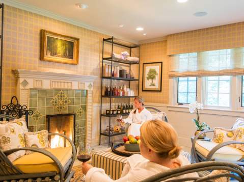 Two ladies in robes sitting in a luxurious sitting room at The Lodge at Glendorn with a lit fireplace. Both women are holding glasses of wine
