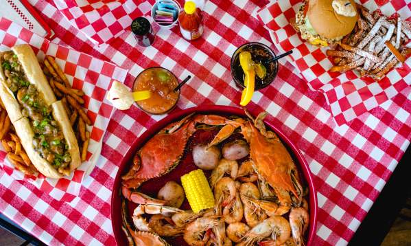 A vibrant seafood platter featuring crabs, shrimp, corn, and potatoes, alongside a mouthwatering hot dog and refreshing drinks on a checkered tablecloth.