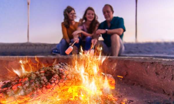 A family of three sitting by a bonfire on the sand roasting marshmallows