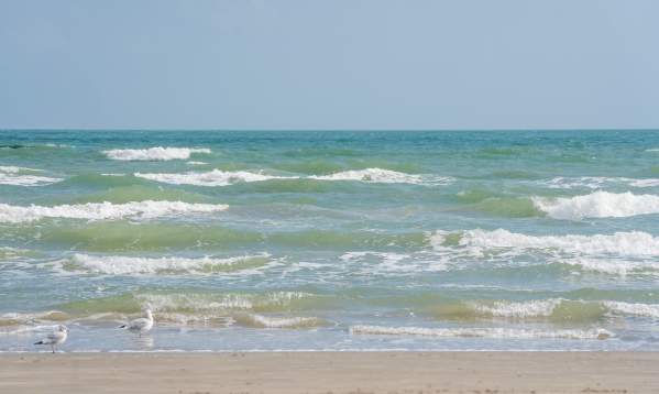 Wide shot of gentle turquoise ocean waves breaking on a sandy beach under a clear blue sky.