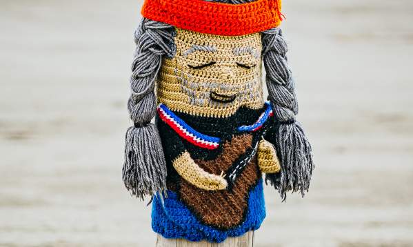 A bollard buddy decorated as Willie Nelson. He has a guitar and his hair in braids with a red headband on.