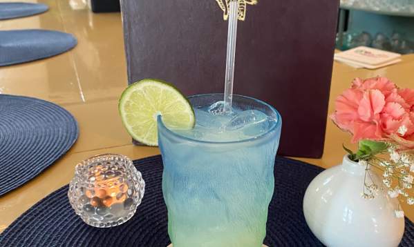 Blue and yellow margarita with a lime on the rim. Flowers and a candle are placed nect to the drink wiht a menu behind it.
