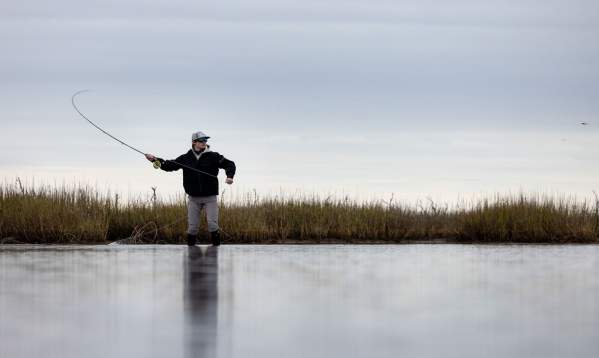 A silhouette of a person fly fishing in a calm marsh at sunset with tall grass in the background.