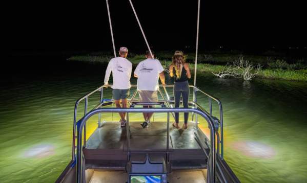 Two men on a boat using bright lights and poles to fish for flounder at night in Port Aransas.