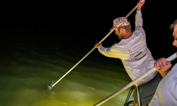 A man in a white long-sleeve shirt uses a pole to gig for flounder from a boat at night.