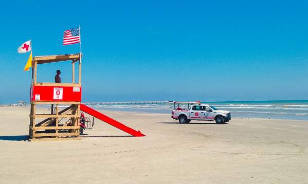 On the beach, a wooden lifeguard tower is painted red across the middle and stairs. Several flags fly from the tower, and a white rescue truck sits near the tower.