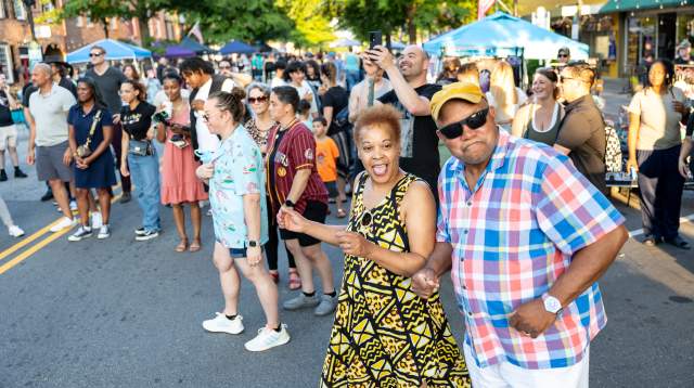 Diverse group of people dancing and smiling at a Fourth Friday event in downtown Fayetteville, NC, with vendor tents and summer sunlight in the background.