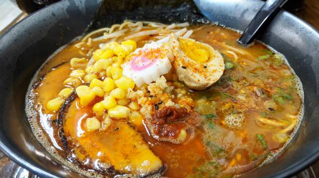 Close-up of a vibrant bowl of miso ramen from Gohan Bistro in Fayetteville, NC, featuring rich broth, sweet corn, marinated egg halves, scallions, noodles, a narutomaki fish cake, and seaweed, all served in a dark ceramic bowl.