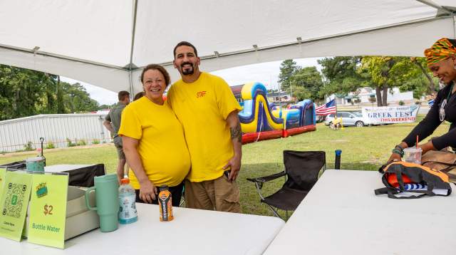 A smiling couple in yellow shirts pose under a white festival tent, with a colorful inflatable obstacle course and Greek Festival banner visible in the background in Fayetteville, NC.