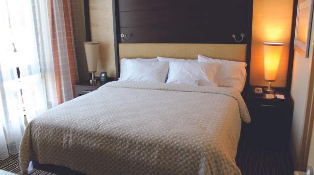 Hotel guest room with a king bed, neutral bedding, bedside tables with lamps, and natural light from a window.