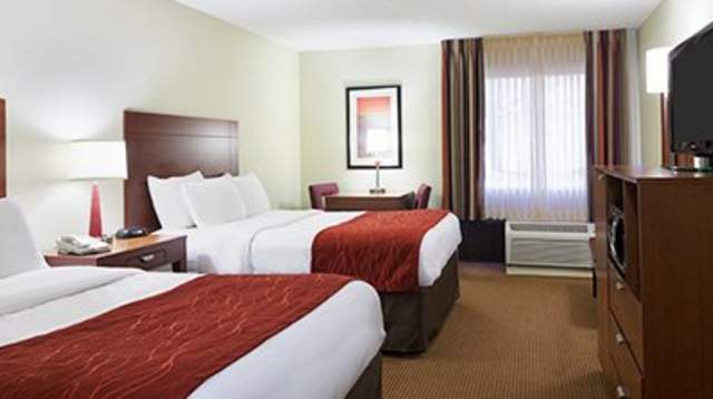 Hotel room with two beds featuring white bedding and red accent throws, wood headboards, bedside tables with lamps, a desk, dresser with TV, and a window with curtains and an in-room air-conditioning unit.