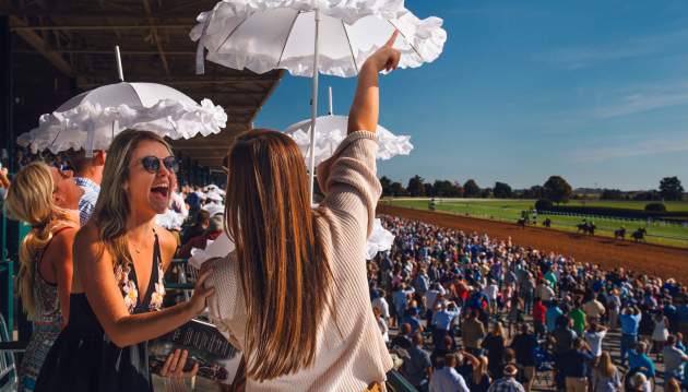 Two young women in a huge crowd at a Keeneland horse race cheer while holding white, lacy parasols.