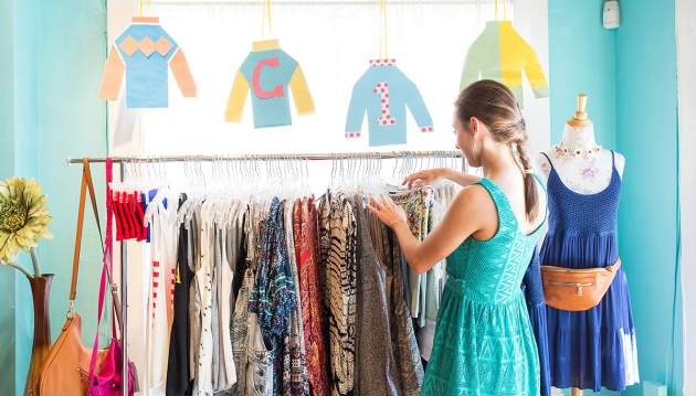 A woman in a teal dress looks through a rack of brightly colored clothing at a shop.