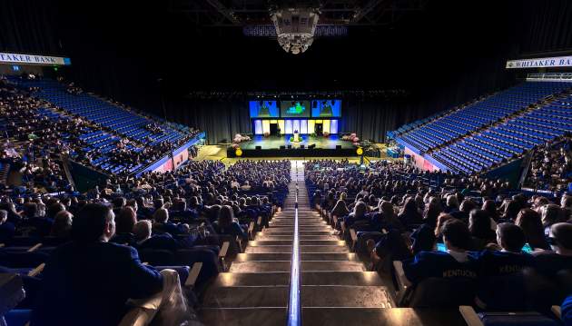 The stadium inside Rupp Arena is filled with blue seats and students wearing University of Kentucky blue while listening to a presenter speak.