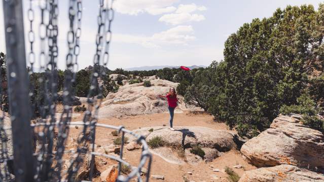 A person dressed in red throwing a disc into a disc golf basket in Three Peaks Recreation Area