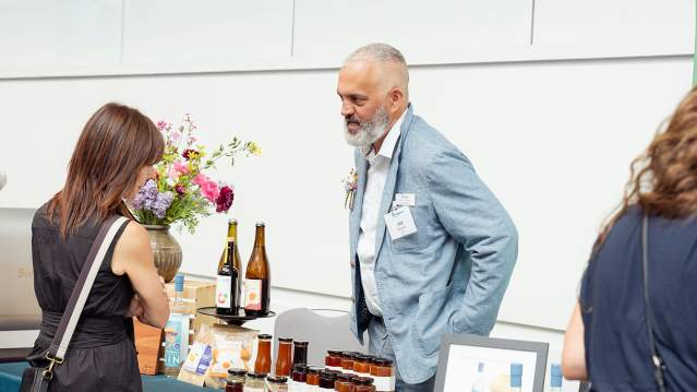 Event table at 2026 Annual Luncheon. Woman talking to booth host. Bottles of wine on booth table.