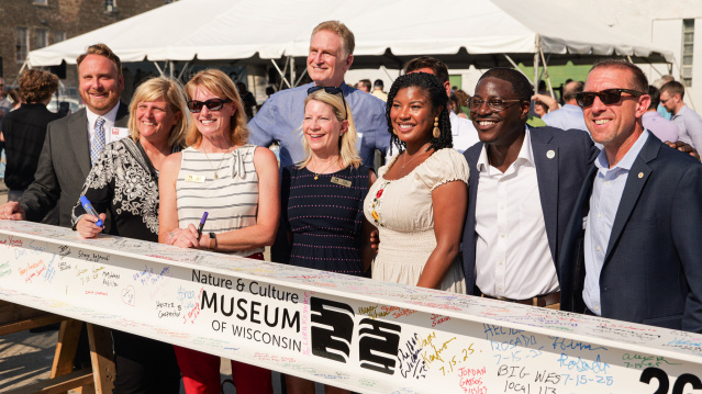 A group of community leaders and attendees stand together outdoors, smiling for a photo behind a large ceremonial ribbon or banner at a public event, with a tent and buildings visible in the background.