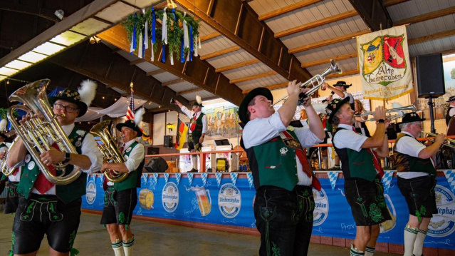 A traditional German band performs indoors at a festival. The musicians are dressed in green vests, white shirts, red ties, and lederhosen. They are playing brass instruments, including a trumpet and tuba. The venue is decorated with festive banners, flags, and blue-and-white streamers hanging from the ceiling. A stage with more performers is visible in the background.