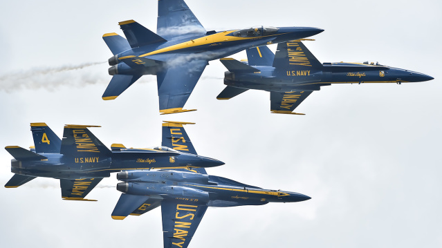 Four U.S. Navy Blue Angels F/A-18 jets perform a close-formation aerial maneuver during an air show, with one jet flying inverted above another. The aircraft are painted in blue and gold with “U.S. NAVY” and “Blue Angels” visible on the fuselage.