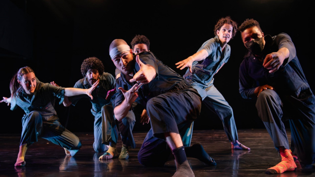 A group of six dancers strike expressive, dynamic poses on stage under dramatic lighting, wearing modern, casual performance outfits.