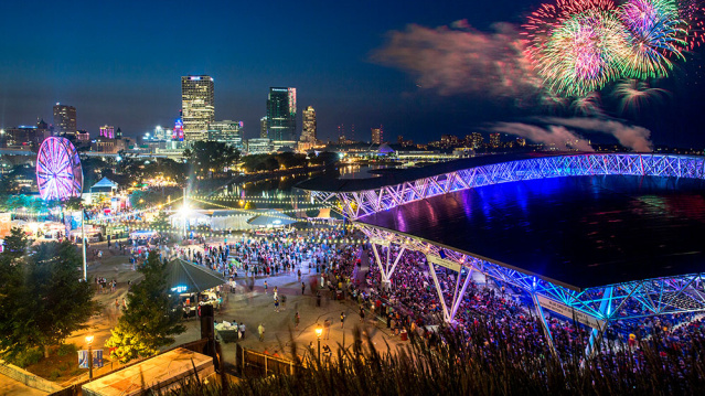 A vibrant nighttime scene at Summerfest grounds in Milwaukee, featuring a large crowd, illuminated Ferris wheel, and the glowing roof of the American Family Insurance Amphitheater. The downtown skyline sparkles in the background as colorful fireworks burst over Lake Michigan, adding to the festive atmosphere.