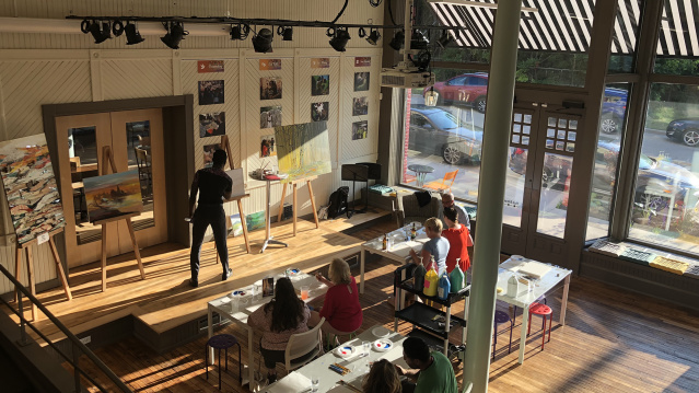 People gather in a bright, open workspace with large windows, high ceilings, and an elevated stage area, participating in a meeting or workshop while natural light fills the room.