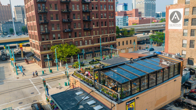 The Lux Skyhaus at Cafe Benelux, a glass greenhouse rooftop dining space in Milwaukee, overlooks a pedestrian plaza with teal lamp posts. The transparent structure reveals lush greenery and diners inside, surrounded by historic red brick buildings and downtown Milwaukee's skyline.