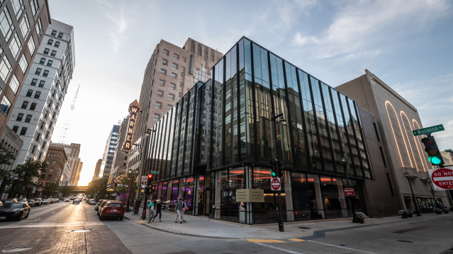 Modern glass-fronted Bradley Symphony Center on a downtown Milwaukee street corner at dusk, reflecting surrounding high-rises; pedestrians cross Wisconsin Avenue while the lit Warner Theatre marquee glows nearby.