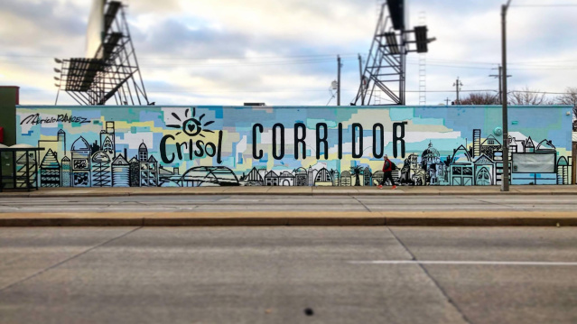 A large outdoor mural reading “Crisol Corridor” with illustrated cityscape elements in shades of blue and yellow. Two pedestrians walk past on the sidewalk in front of the mural under an overcast sky.