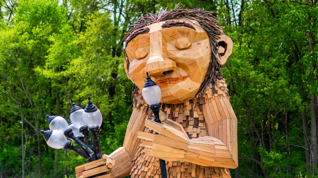 A large wooden sculpture of 'Mama Rosa,' a Thomas Dambo troll installation in Wauwatosa. The friendly giant figure is constructed from reclaimed wood pieces in natural tones, featuring a carved face with a gentle smile and twig-like hair made from bundled branches. The troll holds a bouquet of old-fashioned street lamps in one hand while lifting one lamp to her nose. The sculpture is set against a lush green forest backdrop with dense tree canopy, creating a whimsical scene that blends art with nature.