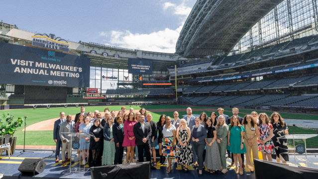 Group photo of attendees at Visit Milwaukee's Annual Meeting, posed on a stage at American Family Field (Milwaukee Brewers stadium) with stadium seating and banners visible in the background.