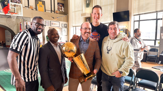 A group of five men pose together at Gee's Clippers. From left to right: Gee in a black and white striped referee shirt, Mayor Johnson in a dark business suit, David Caruso in a brown plaid blazer holding a golden basketball championship trophy,  Steve Novak in a dark shirt, and Peter Feigin in a cream-colored hoodie with a deer logo. The room features basketball memorabilia on the walls including framed jerseys and photos, a basketball hoop, and large windows.