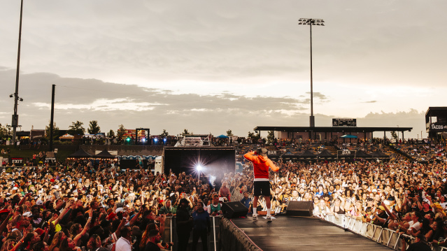 A performer in a red hoodie stands on an outdoor stage facing a massive crowd at sunset. Thousands of fans pack the venue, many holding up phones to capture the moment, while bright stadium lights and food stand signs glow in the background.