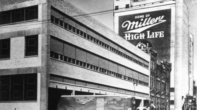 The exterior of Miller Brewing Company with a truck exiting a building. The side of the truck reads, "Miller High Life, The Champagne of Beers." The Miller High Life logo also appears on a much taller building in the background.