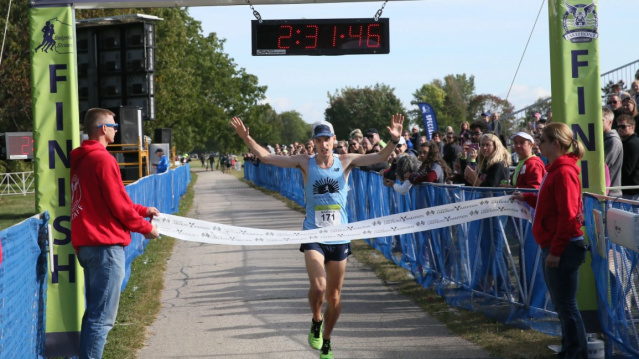 A runner raises their arms in celebration while crossing the finish line at a road race. Spectators line both sides of the course behind barriers, cheering as a race clock overhead displays the finishing time and volunteers hold the finish tape.