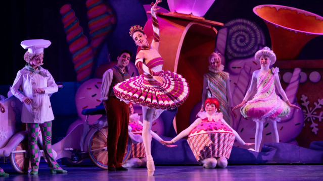 A ballerina in a red-and-white candy-themed costume performs a graceful pose on stage, surrounded by dancers dressed as sweets, including cupcakes and confectioners. The colorful set features oversized candies and whimsical decorations, illuminated by pink and purple stage lighting.